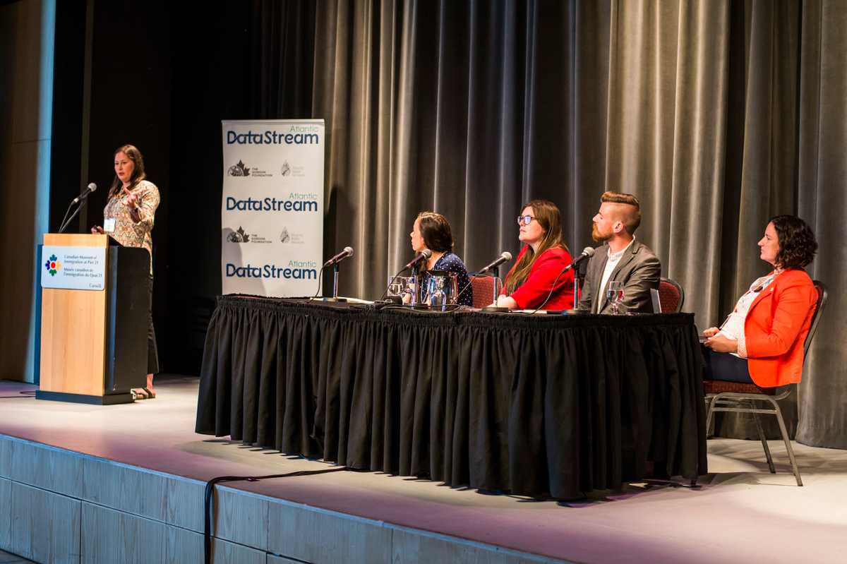 Gila Somers standing at a podium speaking while Carolyn DuBois, Emma Wattie, Graeme Stewart-Robertson, and Elizabeth Hendriks sit at a table on stage